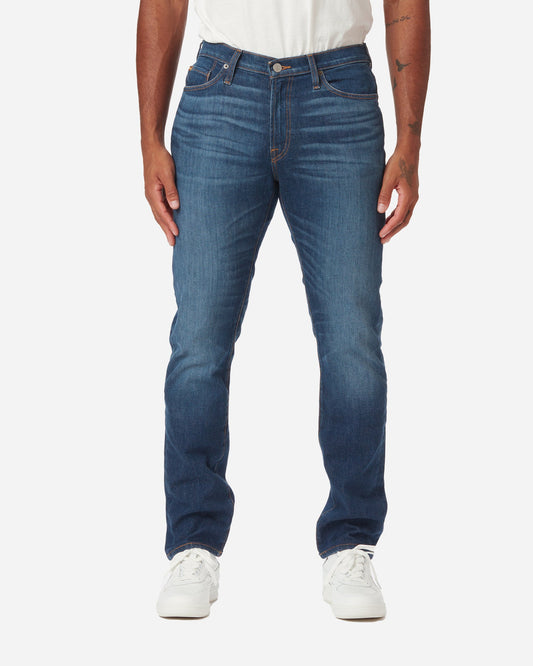 Model facing directly forward wearing a pair of Ace Rivington dark blue athletic taper denim jeans with light stylistic fabric wear on the thigh area with a pair of white Diadora B Elite sneaker shoes and tucked white Ace Rivington super soft supima short-sleeved tee shirt